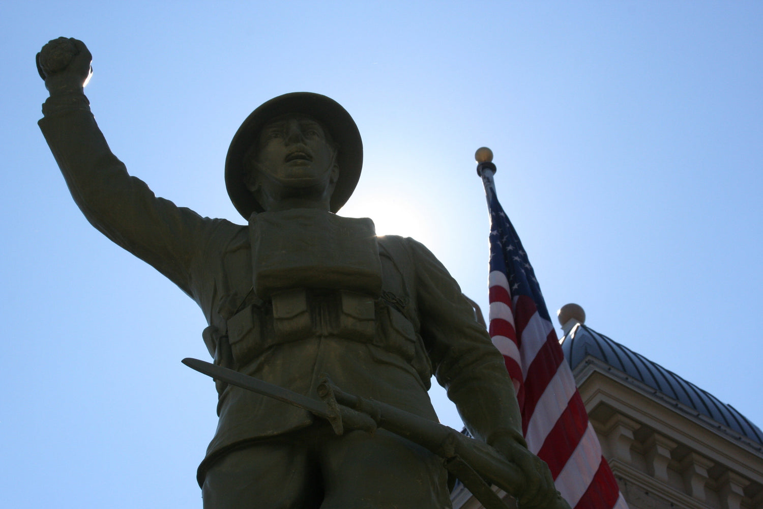WWI Doughboy Soldier Statue backlit by the sun with the American Flag flying in the background